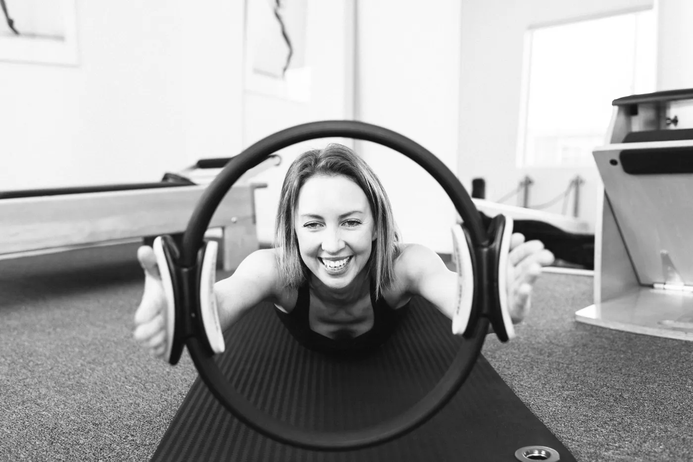 Black and white photo of Emma smiling through a Pilates ring in the studio