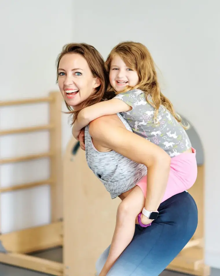 Emma giving her daughter a piggyback ride in her Pilates studio, both laughing